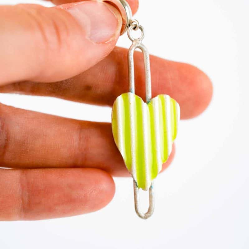 corrugated heart earrings are displayed on a white background. Each earring features a vertically striped enamel heart suspended within a slender sterling silver frame. One pair has bright lime green and white stripes with gold hoops, while the other pair has blue and white stripes with silver hoops. The design is modern and playful, showcasing handcrafted detail and vibrant colour contrast.