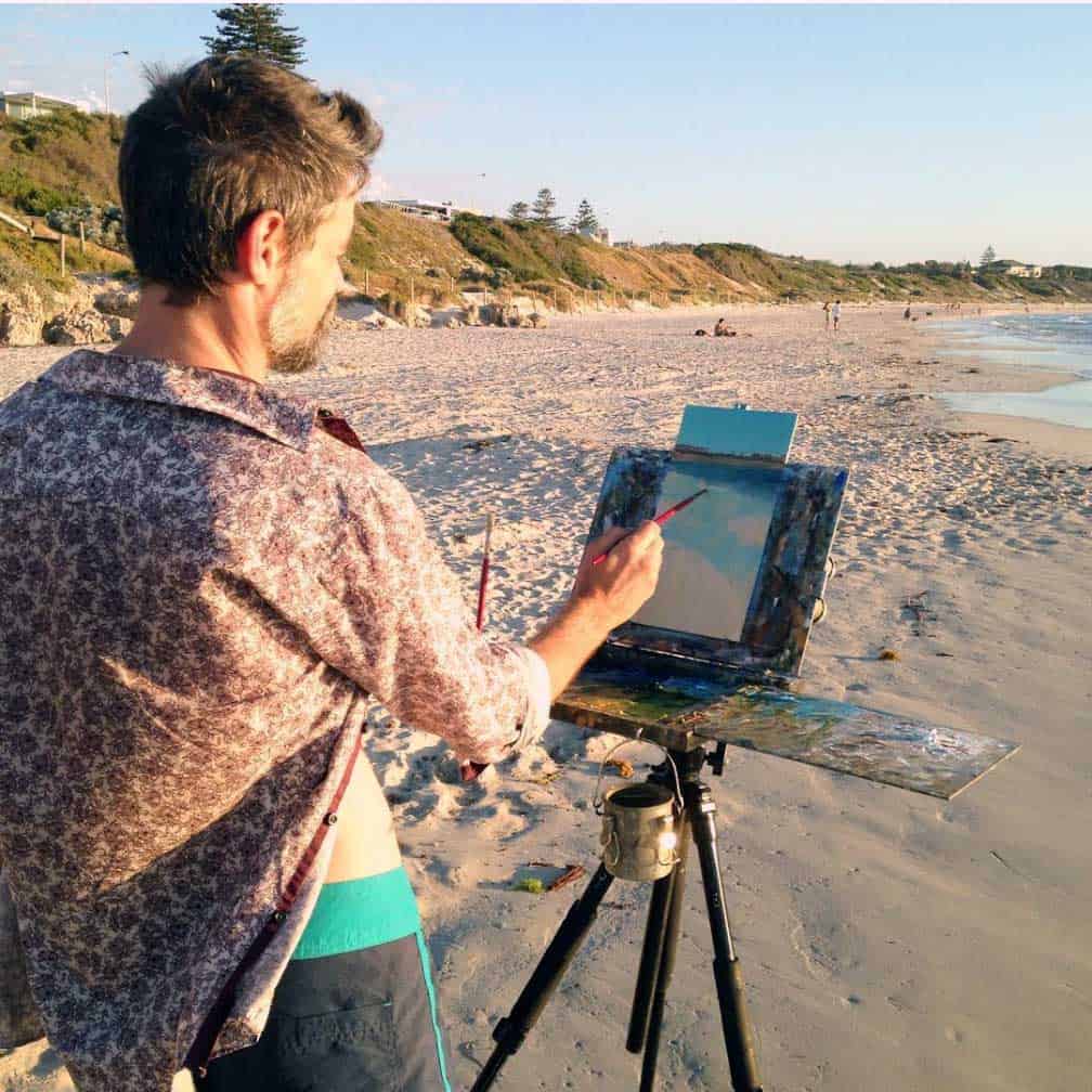 A man with grey hair and a patterned shirt stands on a sandy beach, using a paintbrush to work on a landscape painting fixed to a portable easel tripod.