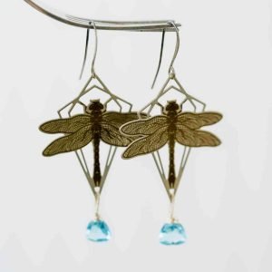 A pair of silver dragonfly earrings hang in front of a plain off-white background. The dragonfly is framed in a geometric diamond shape and has a small blue quartz drop hanging below it