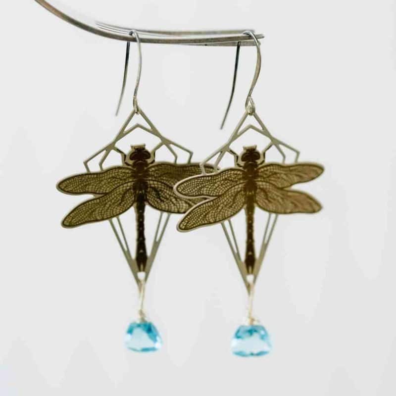 A pair of silver dragonfly earrings hang in front of a plain off-white background. The dragonfly is framed in a geometric diamond shape and has a small blue quartz drop hanging below it
