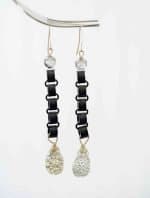 Druzy Agate Earrings (1) A pair of long earrings hang in front of an off-qhite background. The earrings consist of a silver beed follow by a chain of black chains, and end with a large teardrop shaped Druzy Agate. The agate is silver with a rough texture.