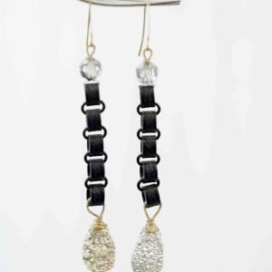 A pair of long earrings hang in front of an off-qhite background. The earrings consist of a silver beed follow by a chain of black chains, and end with a large teardrop shaped Druzy Agate. The agate is silver with a rough texture.