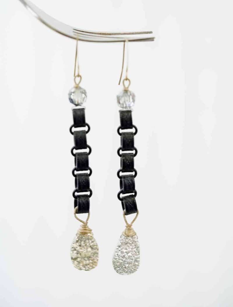 Druzy Agate Earrings (1) A pair of long earrings hang in front of an off-qhite background. The earrings consist of a silver beed follow by a chain of black chains, and end with a large teardrop shaped Druzy Agate. The agate is silver with a rough texture.