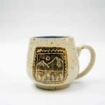 Engraved Mug with Blue Interior (3) A handcrafted ceramic mug with a rounded shape and a single handle, displayed against a plain white background. The mug has a speckled beige glaze and features an engraved design framed by a scalloped border. The design depicts a stylized mountain landscape with a sun or moon above and trees below. The interior of the mug is glazed in a rich blue color, adding a vibrant contrast to the earthy exterior.