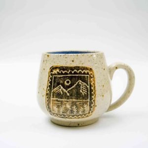 A handcrafted ceramic mug with a rounded shape and a single handle, displayed against a plain white background. The mug has a speckled beige glaze and features an engraved design framed by a scalloped border. The design depicts a stylized mountain landscape with a sun or moon above and trees below. The interior of the mug is glazed in a rich blue color, adding a vibrant contrast to the earthy exterior.