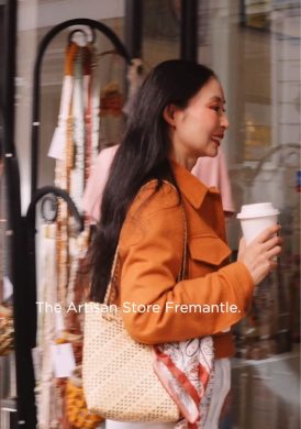 Grace, wearing an orange jacket and carrying a woven bag with a patterned scarf, walks toward The Artisan Store Fremantle holding a takeaway coffee cup. She smiles as she passes displays of handmade jewelry and crafts outside the shop, capturing a warm, lively Fremantle street atmosphere.