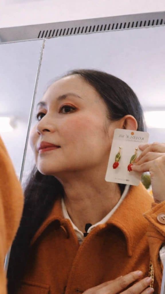 A woman wearing an orange coat looks into a mirror while holding a card with a pair of red and gold earrings beside her ear. Her reflection shows her examining how the earrings might look when worn, with soft indoor lighting and a calm, focused expression.