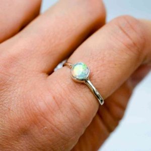A delicate opal shining with yellows and greens is set on a thin sterling silver band. The ring is being worn on the pointer finger of a fair-skinned woman.
