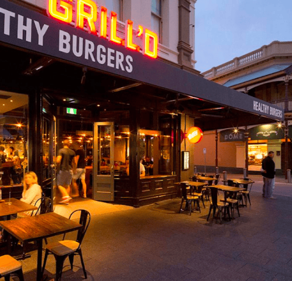 An evening street view of Grill’d Fremantle, a restaurant known for its healthy burgers. The scene shows the warmly lit exterior with a black awning and bright red neon sign reading “GRILL’D.” Several wooden tables and black chairs are arranged outside for diners, with people visible inside and around the entrance. The atmosphere is inviting and lively, with soft lighting and nearby buildings adding to the charm of Fremantle’s café strip.