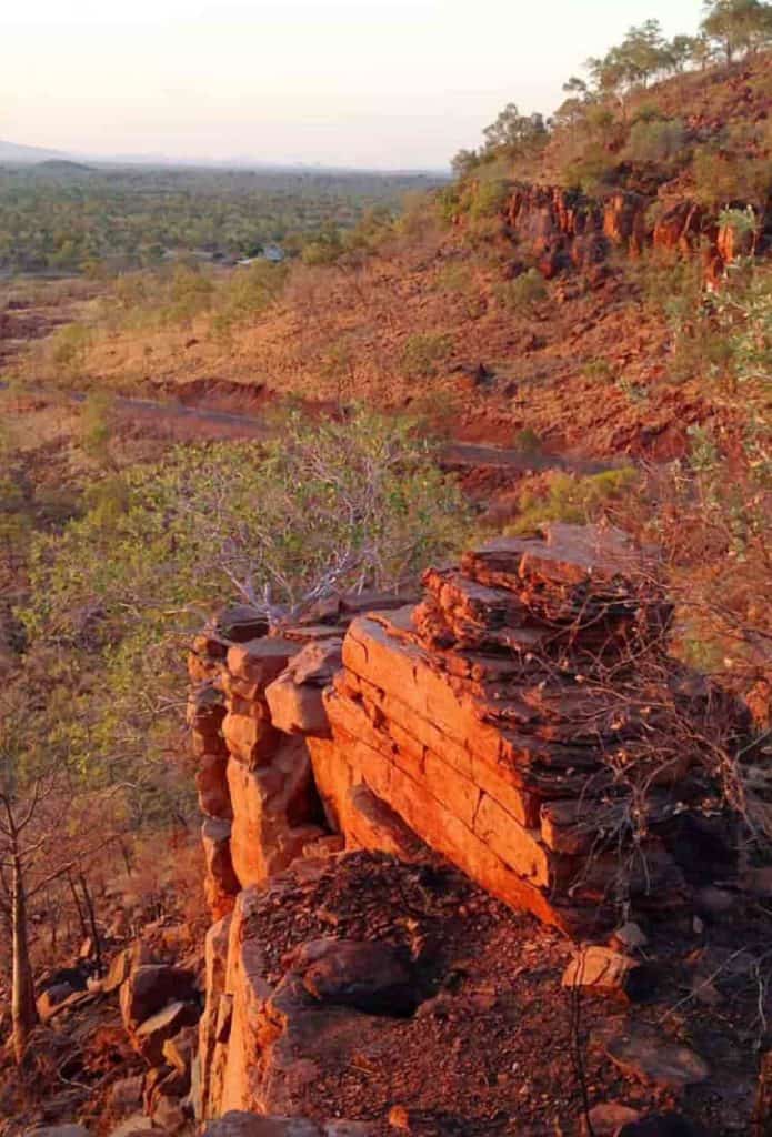 Elevated view of a rugged, red rocky cliff face in Kununurra during sunset, overlooking a vast valley filled with sparse trees and a winding road below.