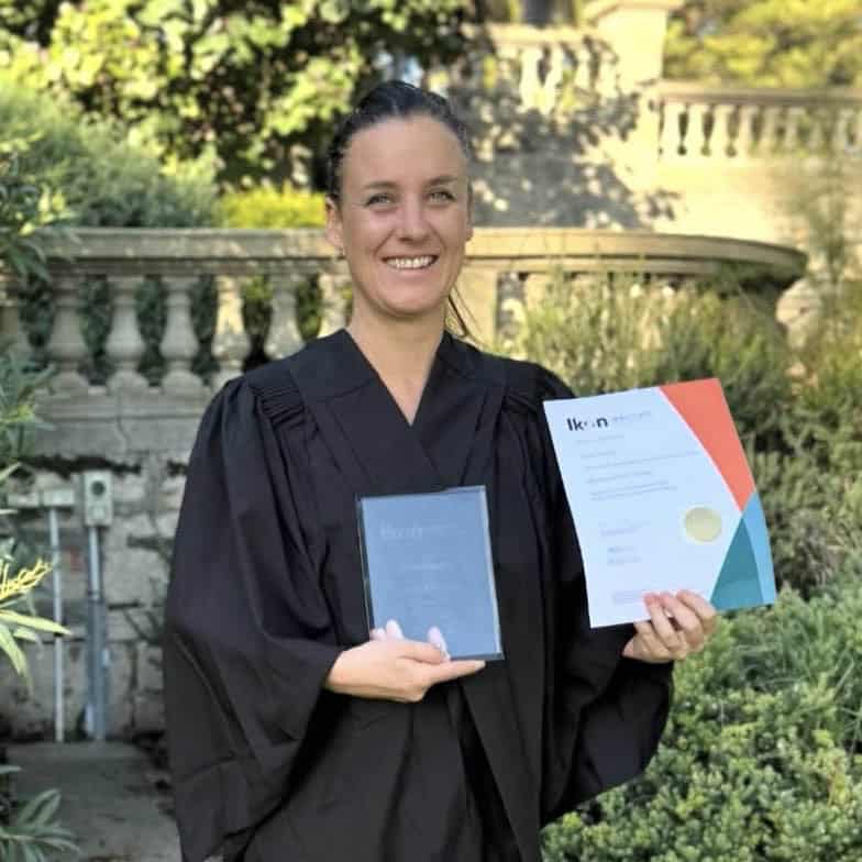 Laura Farnhill stands outdoors in a graduation gown, smiling and holding her Art Therapy degree certificate and an award plaque from Ikon Institute. She is positioned in front of a stone balustrade surrounded by greenery, celebrating her academic achievement.
