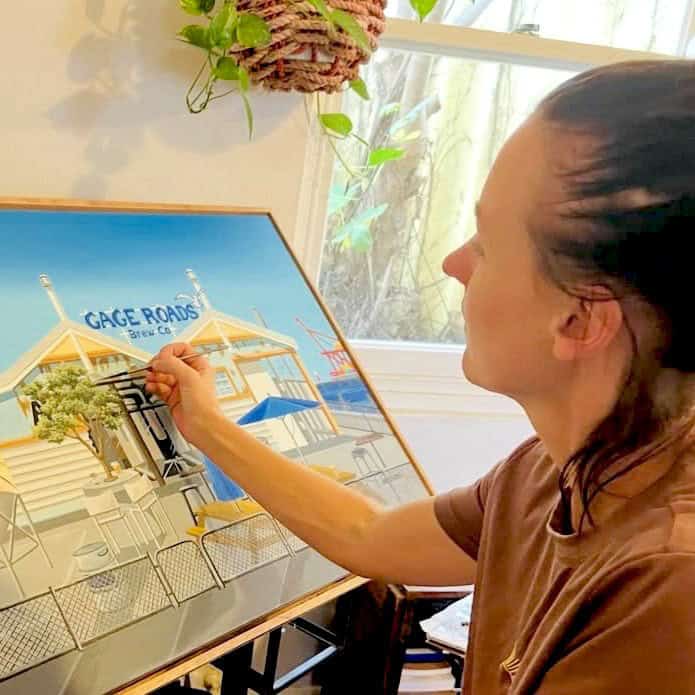 Artist Laura Farnhill sits at a desk painting an acrylic original of Gage Roads Brew Co in Fremantle. The artwork shows the waterfront brewery building with bright blue skies and colourful details. Laura is focused on her work, surrounded by brushes, paints, and art supplies in a cosy studio setting.