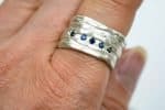 A close-up of a handcrafted sterling silver ring worn on a finger. The ring features a textured, layered Mitsuro-style design with five small round blue sapphires set in a horizontal line across the centre. The silver surface has an organic, rippled finish that catches the light, highlighting the deep blue sparkle of the gemstones.