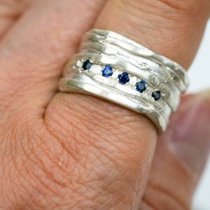A close-up of a handcrafted sterling silver ring worn on a finger. The ring features a textured, layered Mitsuro-style design with five small round blue sapphires set in a horizontal line across the centre. The silver surface has an organic, rippled finish that catches the light, highlighting the deep blue sparkle of the gemstones.
