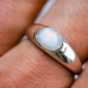 A white opal set into a thick Sterling silver band is worn on the middle finger of a fair-skinned woman. The photo is very zoomed in.