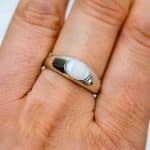 The opal ring is worn on the ring finger of a light-skinned woman. The photo is close cropped and only thee base and first knuckle can be seen. The ring sits between them