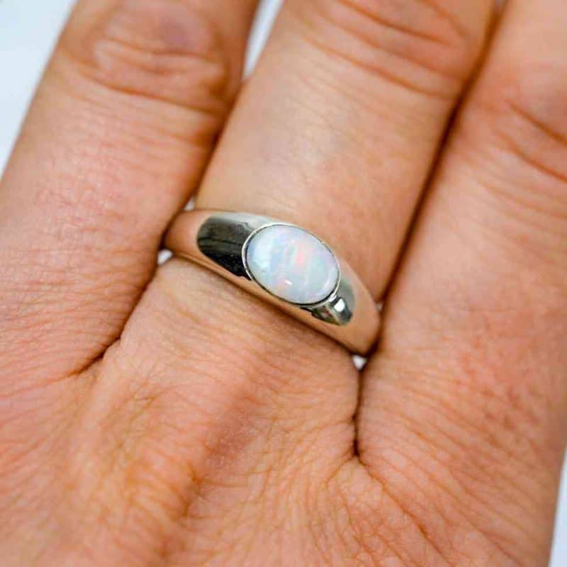 The opal ring is worn on the ring finger of a light-skinned woman. The photo is close cropped and only thee base and first knuckle can be seen. The ring sits between them