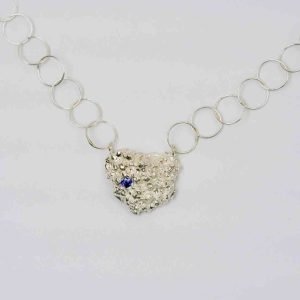 A close up image of a silver necklace pendant. The pendant is triangular in shape and shows the rough texture of silver wax-cast coral. On the left side of the pendant a small dark blue sapphire is set into the silver. The pendant is attached on either end to a chain made of small, thin silver rings