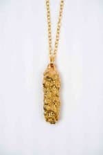 Neo Now Jewellery -6 An obling pendant made of bronze hand on a thin golden chain infront of an off-white background. The pendant is made from a maleluca seedpod cast in bronze
