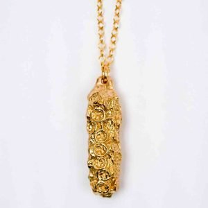 An obling pendant made of bronze hand on a thin golden chain infront of an off-white background. The pendant is made from a maleluca seedpod cast in bronze