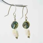 Pearl and Paua Earrings (1) A close-up image of a pair of handmade dangle earrings hanging from a metallic display. Each earring features a round paua bead with iridescent shades of green, blue, and violet, paired with a rectangular white freshwater pearl below. The earrings are finished with small silver beads at the base and simple curved hooks, highlighting their natural, ocean-inspired design against a soft, neutral background.