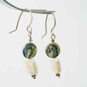 A close-up image of a pair of handmade dangle earrings hanging from a metallic display. Each earring features a round paua bead with iridescent shades of green, blue, and violet, paired with a rectangular white freshwater pearl below. The earrings are finished with small silver beads at the base and simple curved hooks, highlighting their natural, ocean-inspired design against a soft, neutral background.
