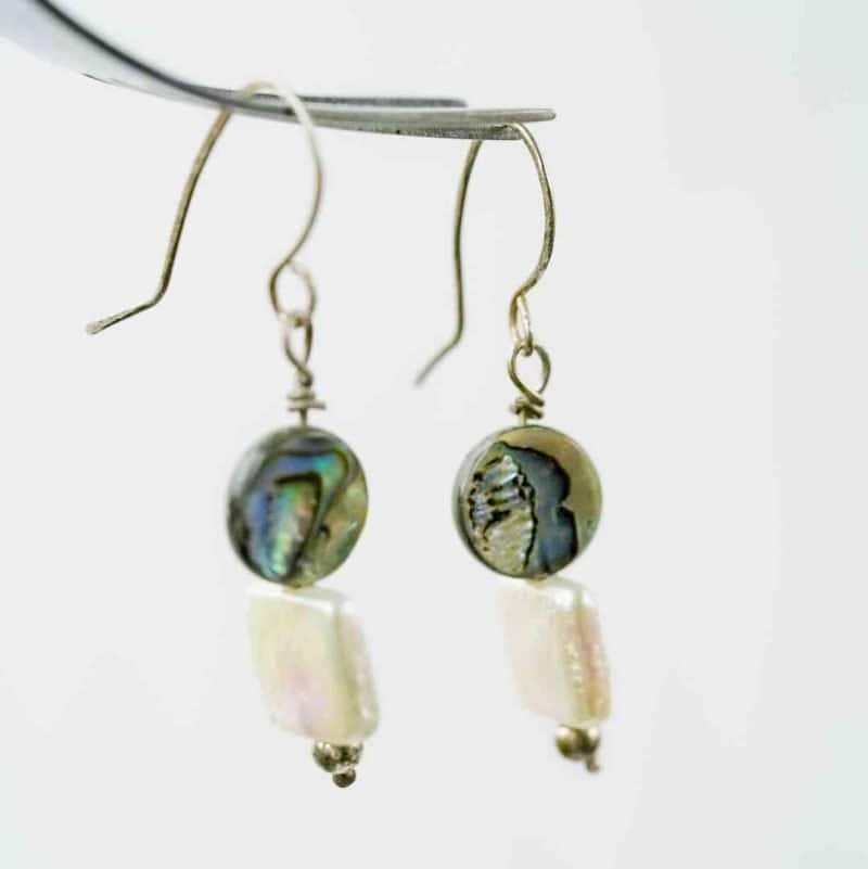 Pearl and Paua Earrings (1) A close-up image of a pair of handmade dangle earrings hanging from a metallic display. Each earring features a round paua bead with iridescent shades of green, blue, and violet, paired with a rectangular white freshwater pearl below. The earrings are finished with small silver beads at the base and simple curved hooks, highlighting their natural, ocean-inspired design against a soft, neutral background.