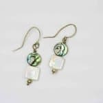 Pearl and Paua Earrings (2) A pair of handmade dangle earrings displayed on a light grey background. Each earring features a round abalone shell bead with iridescent shades of green, blue, and violet, paired with a square white freshwater pearl below. The earrings are finished with small silver beads at the base and simple curved hooks, highlighting their natural, ocean-inspired design and artisan craftsmanship