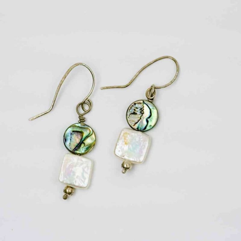 A pair of handmade dangle earrings displayed on a light grey background. Each earring features a round abalone shell bead with iridescent shades of green, blue, and violet, paired with a square white freshwater pearl below. The earrings are finished with small silver beads at the base and simple curved hooks, highlighting their natural, ocean-inspired design and artisan craftsmanship