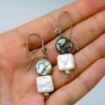 Pearl and Paua Earrings (3) A close-up of a hand holding a pair of handmade dangle earrings. Each earring features a round abalone shell bead with iridescent shades of green, blue, and violet, paired with a square white freshwater pearl below. The earrings are finished with small silver beads and simple curved hooks, highlighting their natural ocean-inspired design and artisan craftsmanship against a soft neutral background.
