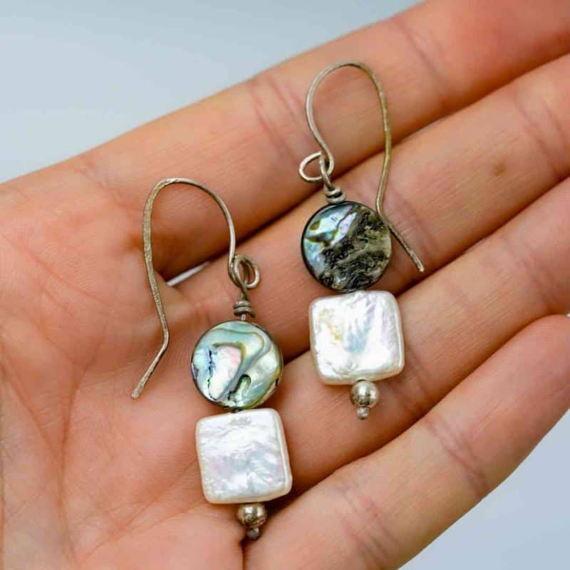 A close-up of a hand holding a pair of handmade dangle earrings. Each earring features a round abalone shell bead with iridescent shades of green, blue, and violet, paired with a square white freshwater pearl below. The earrings are finished with small silver beads and simple curved hooks, highlighting their natural ocean-inspired design and artisan craftsmanship against a soft neutral background.