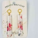 Pink flower drop earrings (1) A pair of long rectangular earrins made of polymer clay with a pink floral display. The earrings are attached to a gold hoop at the top of the polymer clay, The earrings are displayed on a piece of card with the words 'Pretty Mili Clay' at the top in a curve.