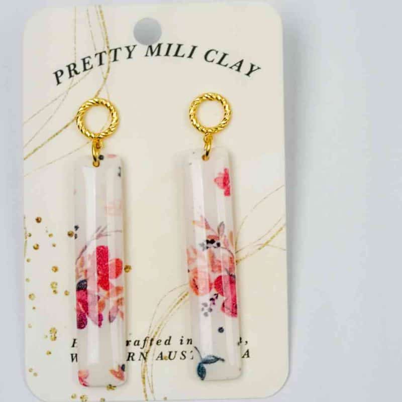 Pink flower drop earrings (1) A pair of long rectangular earrins made of polymer clay with a pink floral display. The earrings are attached to a gold hoop at the top of the polymer clay, The earrings are displayed on a piece of card with the words 'Pretty Mili Clay' at the top in a curve.