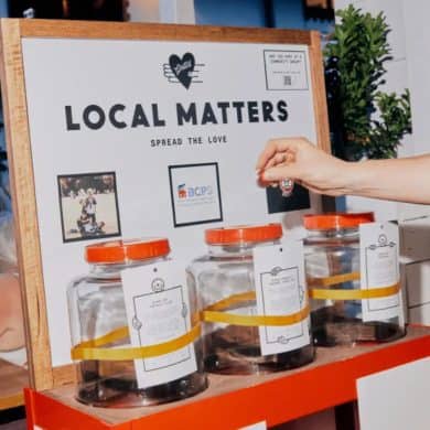A close-up photo of Grill’d Fremantle’s Local Matters donation display. The setup features three large clear jars with orange lids, each labeled with a local community group’s description. A hand is seen placing a token into one of the jars. The background sign reads “Local Matters – Spread the Love” with the Grill’d logo above and smaller logos and photos representing participating organisations. The scene captures the restaurant’s community initiative encouraging customers to support local causes.