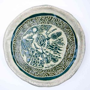 Handcrafted ceramic plate featuring intricate sgraffito carvings of Australian bush flowers surrounded by a decorative wave‑patterned border. The plate has a natural speckled glaze with deep green detailing and a slightly irregular rim, highlighting its artisanal, handmade character.