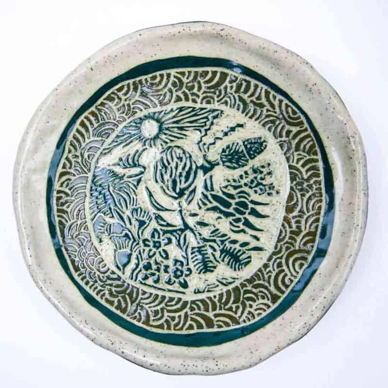Handcrafted ceramic plate featuring intricate sgraffito carvings of Australian bush flowers surrounded by a decorative wave‑patterned border. The plate has a natural speckled glaze with deep green detailing and a slightly irregular rim, highlighting its artisanal, handmade character.