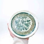 A hand holds a small, handcrafted ceramic plate featuring intricate sgraffito carvings of Australian bush flowers. The design reveals deep green glaze beneath a creamy surface, framed by a decorative wave‑patterned border and a slightly irregular rim. The background is bright and minimal, emphasizing the plate’s detailed artistry and organic texture.