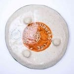 The underside of a handmade ceramic plate with a speckled beige glaze and a central terracotta circle. The circle features an engraved geometric symbol and the words “SEEDS GROW FLOWERS… LIFE” carved around it. A round studio label reading “Amanda Forward Studio 31” is visible on the left side. The surface has three raised bumps, adding texture and character to the piece.