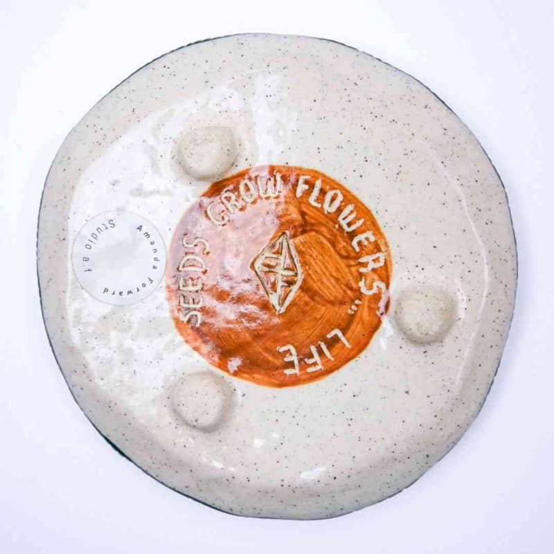 The underside of a handmade ceramic plate with a speckled beige glaze and a central terracotta circle. The circle features an engraved geometric symbol and the words “SEEDS GROW FLOWERS… LIFE” carved around it. A round studio label reading “Amanda Forward Studio 31” is visible on the left side. The surface has three raised bumps, adding texture and character to the piece.