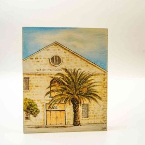 A pyrography image of the WA Shiprewck Museum burned into light timber sits on a white background. The sky is clear and blue, and the palm tree in front of the museum appears to blow in the wind.