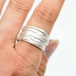 A close-up of a hand wearing a handcrafted sterling silver ring with a layered, textured design. The ring features multiple overlapping bands that create an organic, rippled surface, showcasing the distinctive Mitsuro Hikime technique. The silver has a soft matte finish, highlighting its artisanal craftsmanship and sculptural form.