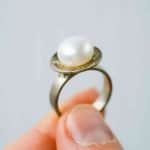Sterling Silver and Pearl ring (3) A close-up of a hand holding a silver ring featuring a single white pearl set on a flat circular base. The pearl has a smooth, lustrous surface, and the background is a soft, neutral grey that highlights the ring’s details.
