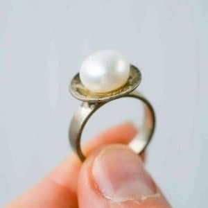 A close-up of a hand holding a silver ring featuring a single white pearl set on a flat circular base. The pearl has a smooth, lustrous surface, and the background is a soft, neutral grey that highlights the ring’s details.