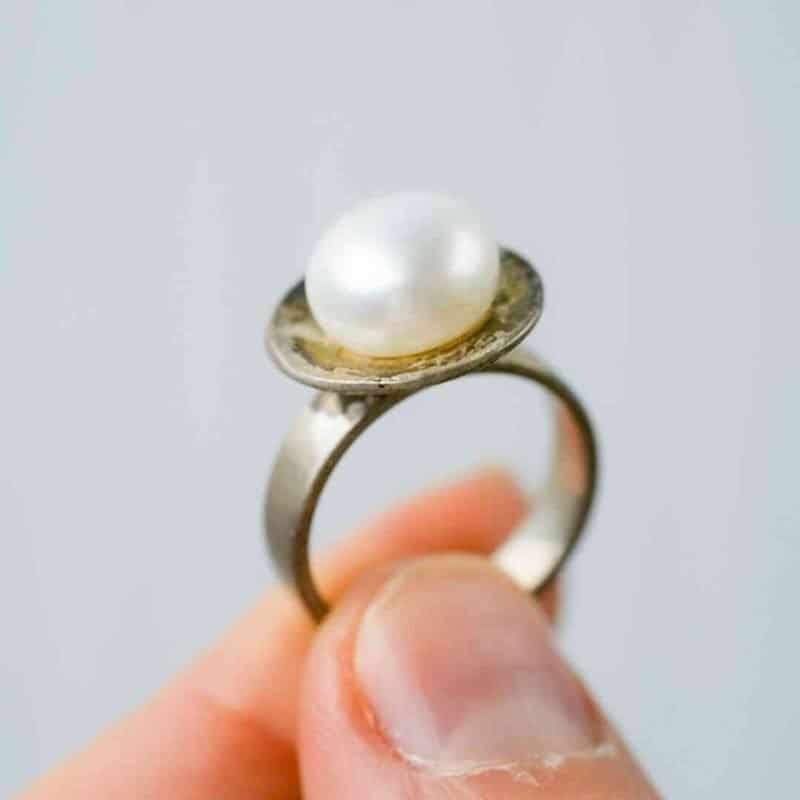 Sterling Silver and Pearl ring (3) A close-up of a hand holding a silver ring featuring a single white pearl set on a flat circular base. The pearl has a smooth, lustrous surface, and the background is a soft, neutral grey that highlights the ring’s details.