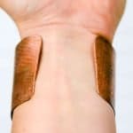 Textured Copper Cuff (1) A close-up of a wrist wearing a wide, handcrafted copper cuff bracelet. The cuff has a warm reddish-brown tone with a natural patina and subtle hammered texture, showing variations in color and surface marks. Its open-ended design curves gently around the wrist, highlighting the artisanal craftsmanship and organic finish against a soft, neutral background.