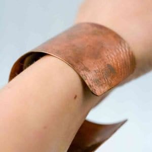 A close-up of a wrist wearing a wide, handcrafted copper cuff bracelet. The cuff has a natural reddish-brown tone with visible texture and patina, showing subtle variations in color and surface marks. Its open-ended design curves gently around the wrist, highlighting the organic, artisanal quality of the metalwork against a soft, neutral background.