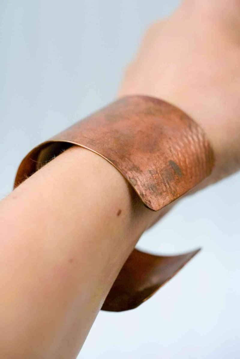 Textured Copper Cuff (4) A close-up of a wrist wearing a wide, handcrafted copper cuff bracelet. The cuff has a natural reddish-brown tone with visible texture and patina, showing subtle variations in color and surface marks. Its open-ended design curves gently around the wrist, highlighting the organic, artisanal quality of the metalwork against a soft, neutral background.