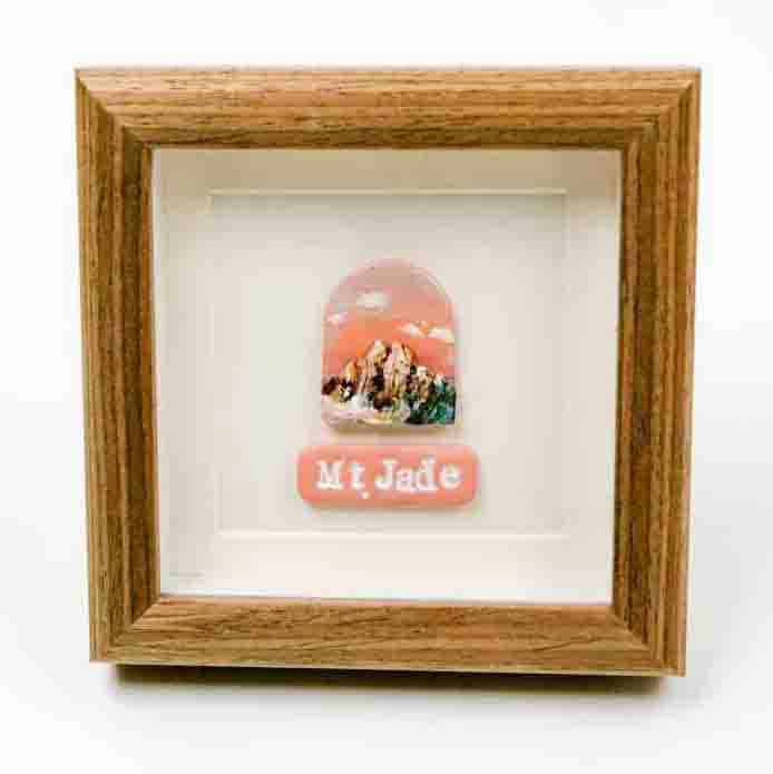 Winimum Handmade -Mt Jade framed A small framed artwork featuring a depiction of Mt. Jade, with rugged peaks under a warm pink and orange sky. Below the illustration, a matching pink plaque displays the words “Mt. Jade” in white lettering. The piece is set within a wooden frame against a white background, creating a clean and elegant presentation.