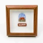 A small framed artwork featuring a depiction of Uluru, the iconic red sandstone monolith in central Australia, under a blue sky with scattered clouds. Below the illustration, a brown plaque displays the word “Uluru” in white lettering. The piece is set within a wooden frame against a white background, creating a clean, minimalist presentation.