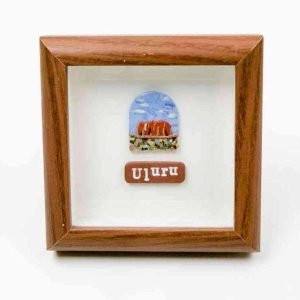 A small framed artwork featuring a depiction of Uluru, the iconic red sandstone monolith in central Australia, under a blue sky with scattered clouds. Below the illustration, a brown plaque displays the word “Uluru” in white lettering. The piece is set within a wooden frame against a white background, creating a clean, minimalist presentation.